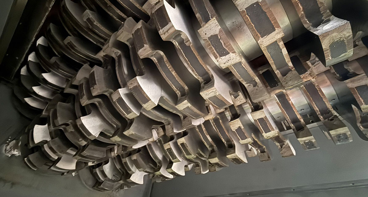 Cutter shaft layout inside the Geording four-shaft shredder: two primary cutting shafts intermesh with two upper clearance shafts above an interchangeable discharge screen — the geometry that drives automatic recirculation until material reaches the target particle size.